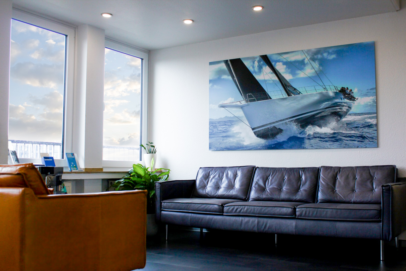 Wartezimmer vor der Anmeldung mit offener Fläche, eine Schwarzen breiten Sofa, einem braunen massiven Sessel und einem großen Bild einer Yacht